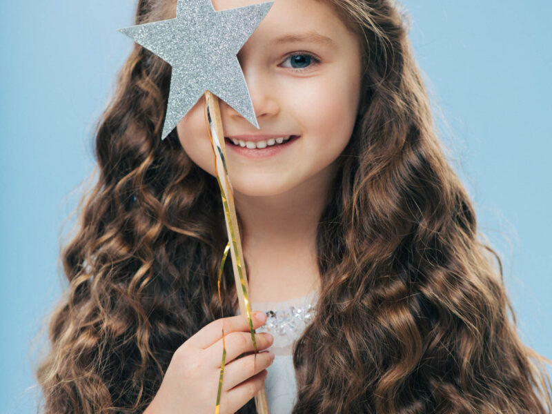 Isolated image of satisfied beautiful curly woman with blue eyes, covers face with magic wand, has long curly hair, poses over blue background, gentle smile on face, makes wish, dreams about something