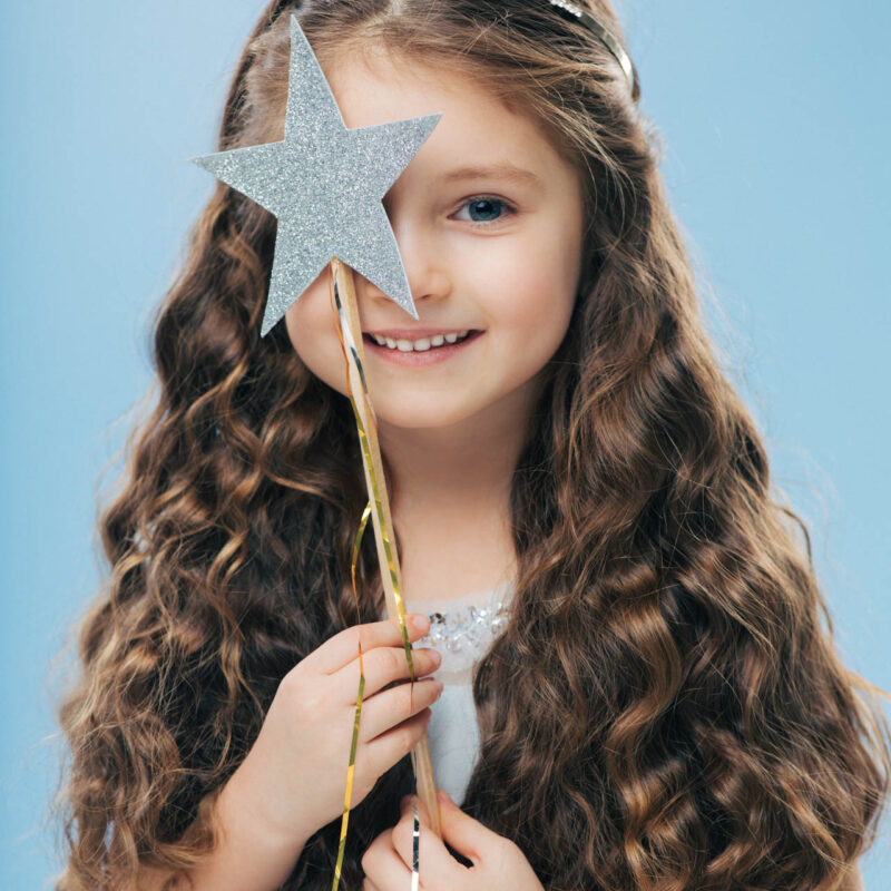 Isolated image of satisfied beautiful curly woman with blue eyes, covers face with magic wand, has long curly hair, poses over blue background, gentle smile on face, makes wish, dreams about something